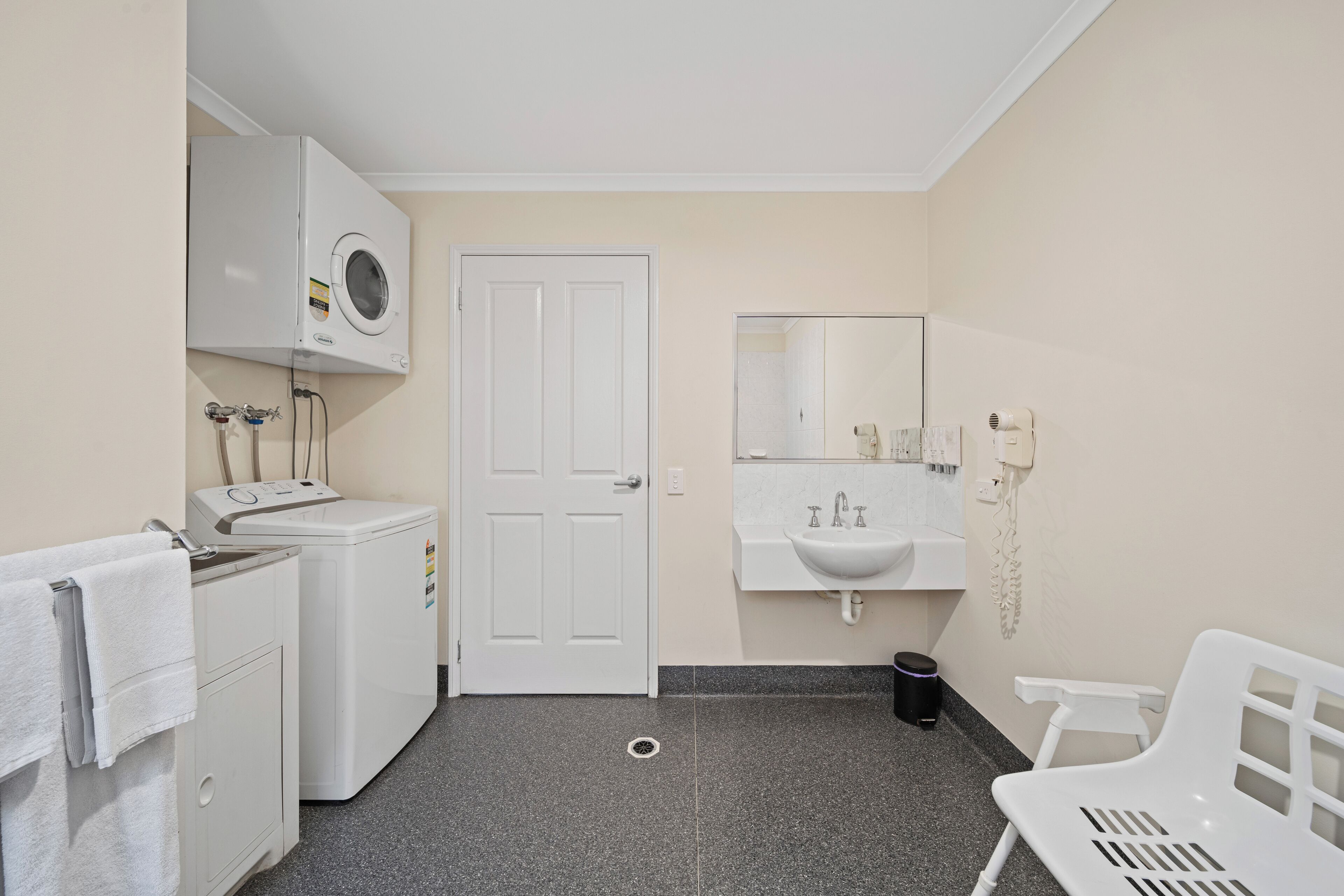 A small, clean bathroom with a washing machine, a sink, a mirror, and a white chair.