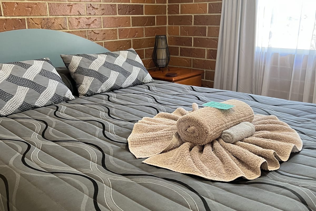 A neatly made bed with a patterned bedspread and two pillows, a small table with a lamp, and a brick wall.