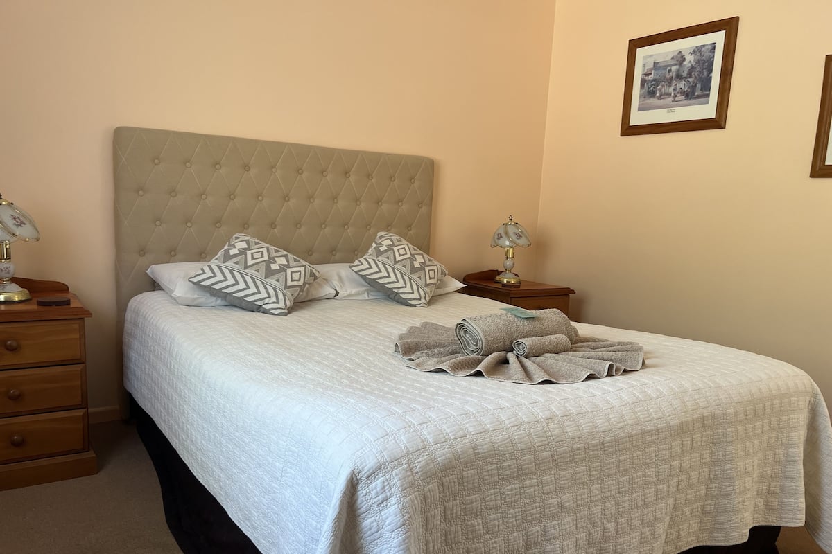 A neatly made bed with a quilted bedspread, two patterned pillows, and a rolled towel on the bed. There are two bedside tables with lamps and a framed picture on the wall.