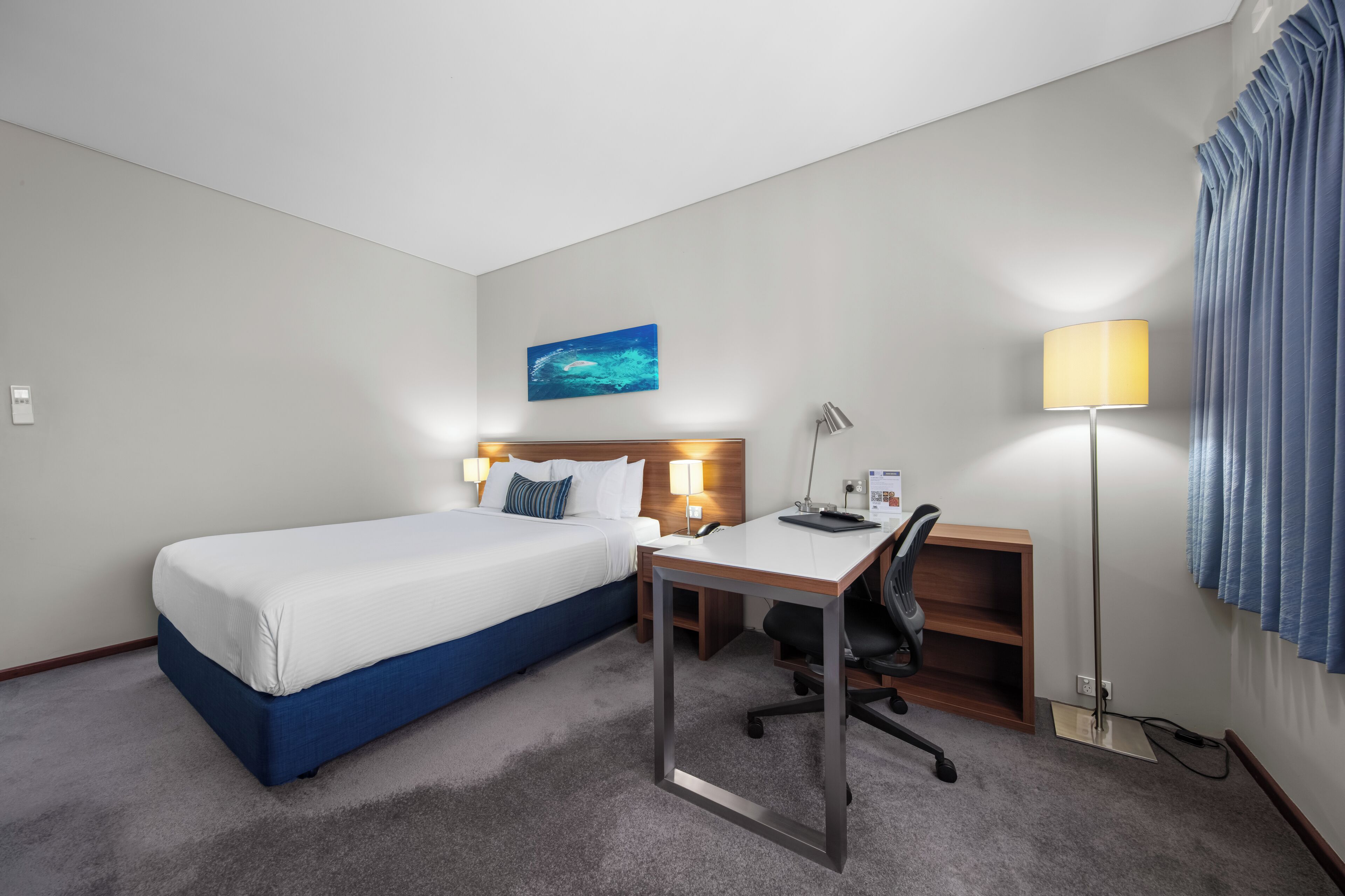 Una habitación de hotel con una cama individual, un escritorio con silla, una lámpara de pie y un cuadro en la pared.