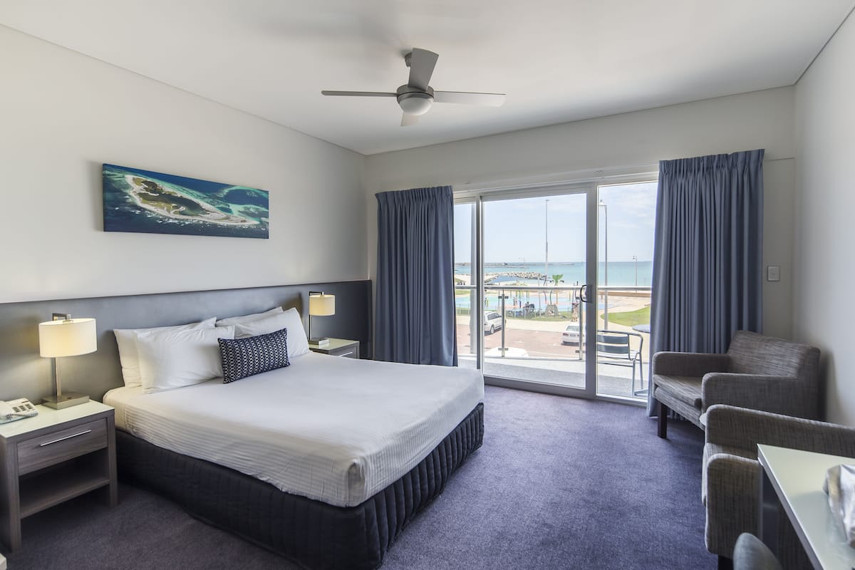 A hotel room with a bed, a desk, a chair, a nightstand, a lamp, a ceiling fan, and a large window with curtains.