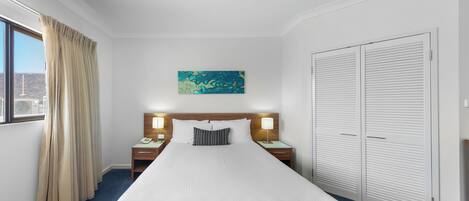 Habitación de hotel con una cama grande, dos mesitas de noche con lámparas, una mesita auxiliar, una ventana con cortinas, una obra de arte colgada en la pared y un conjunto de armarios blancos.