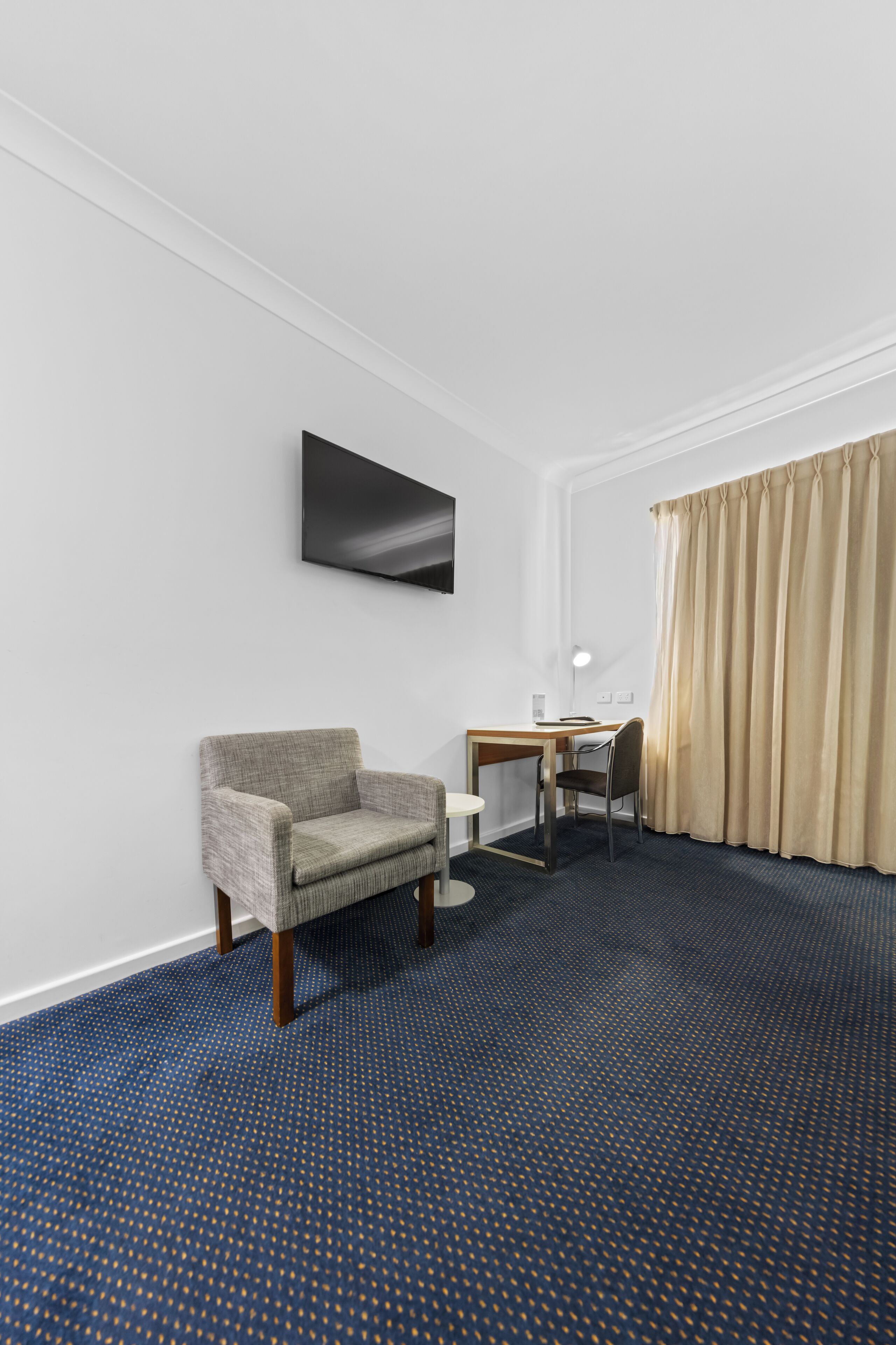 A hotel room with a blue carpet, a grey armchair, a wooden desk, a flat-screen TV mounted on the wall, and a window with beige curtains.