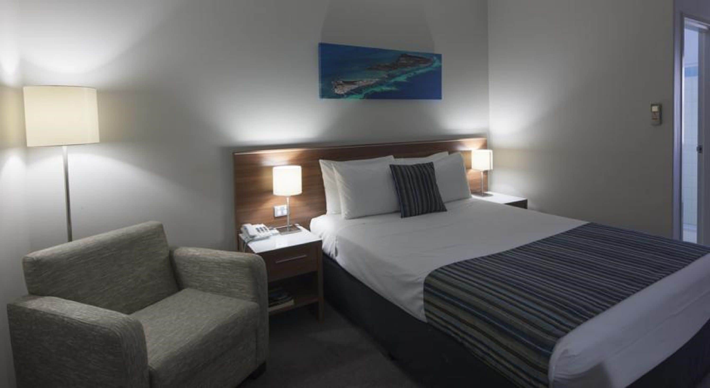 Une chambre d’hôtel avec un lit, une chaise, une table de chevet avec une lampe et un tableau au mur.