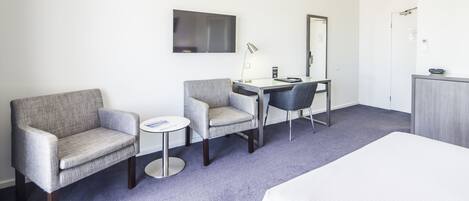 A hotel room with a bed, two armchairs, a small round table, a desk with a chair, a flat-screen TV, a mirror, and a wardrobe.