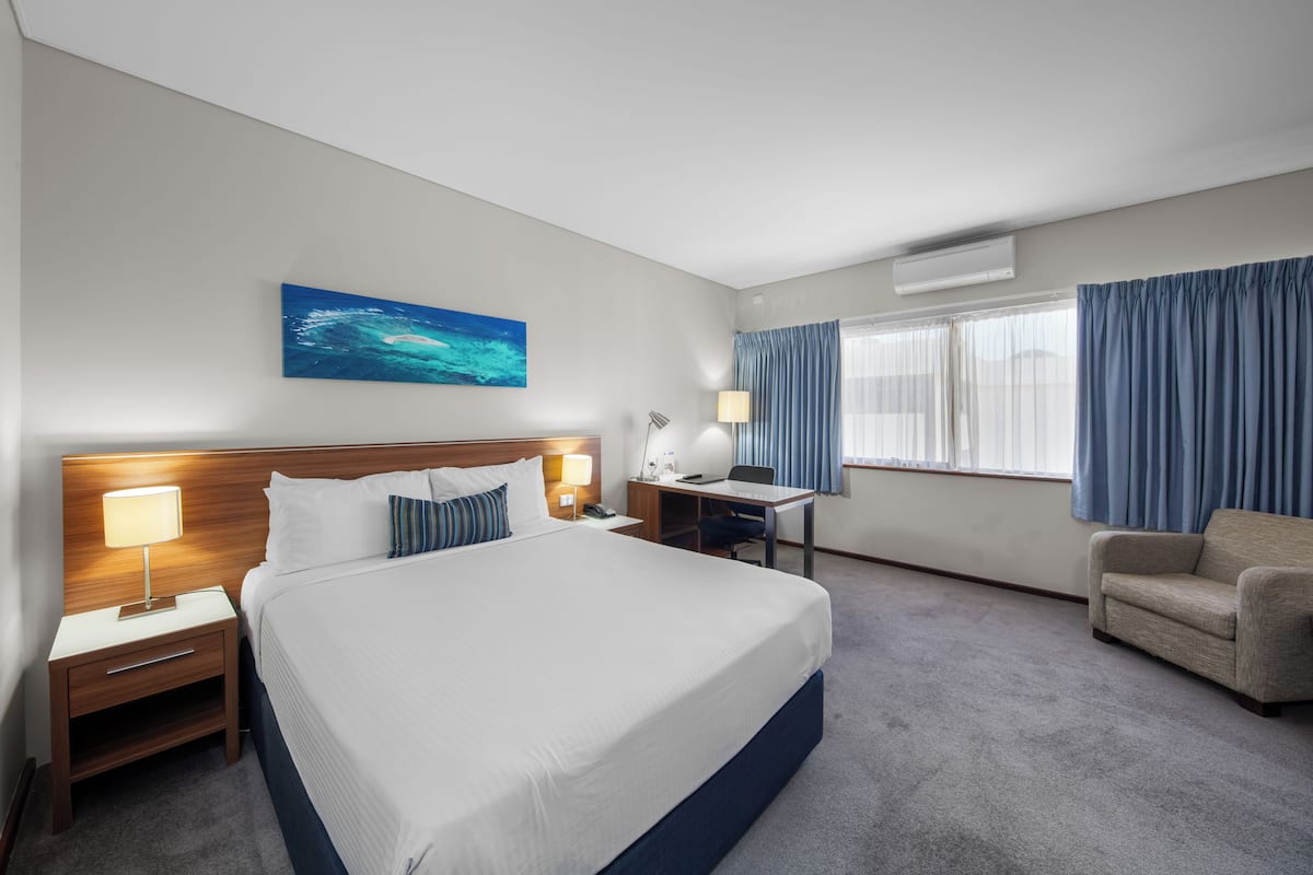 A hotel room with a large bed, a desk, a chair, a nightstand, a lamp, a painting, and a window with curtains.