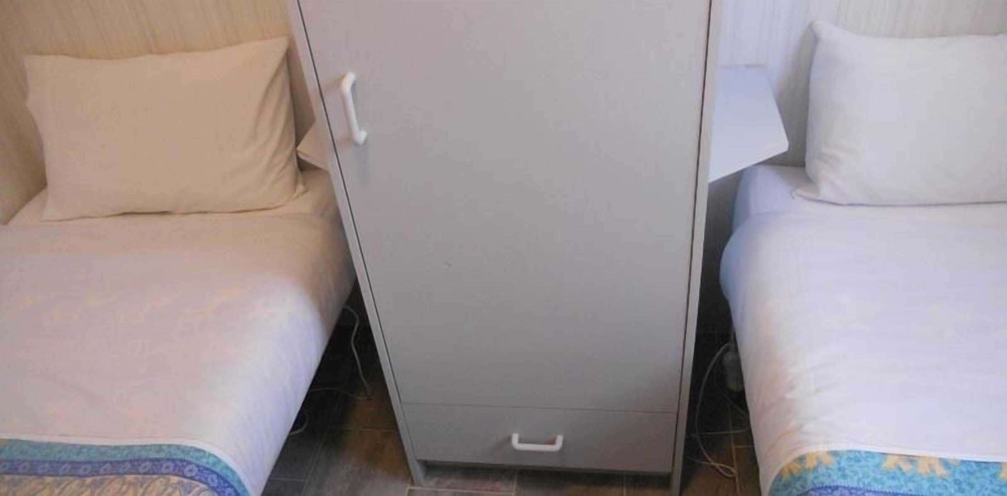 A room with two beds, each with a white mattress and a pillow. There is a grey wardrobe in the center of the room.