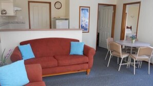 Una sala de estar con un sofá rojo, cojines azules, una mesa de comedor con cuatro sillas, un reloj y una puerta que da a otra habitación.