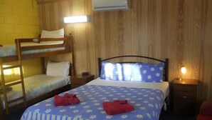 Una habitación con una cama litera, una cama individual, paredes de madera y una mesita de noche con lámpara.
