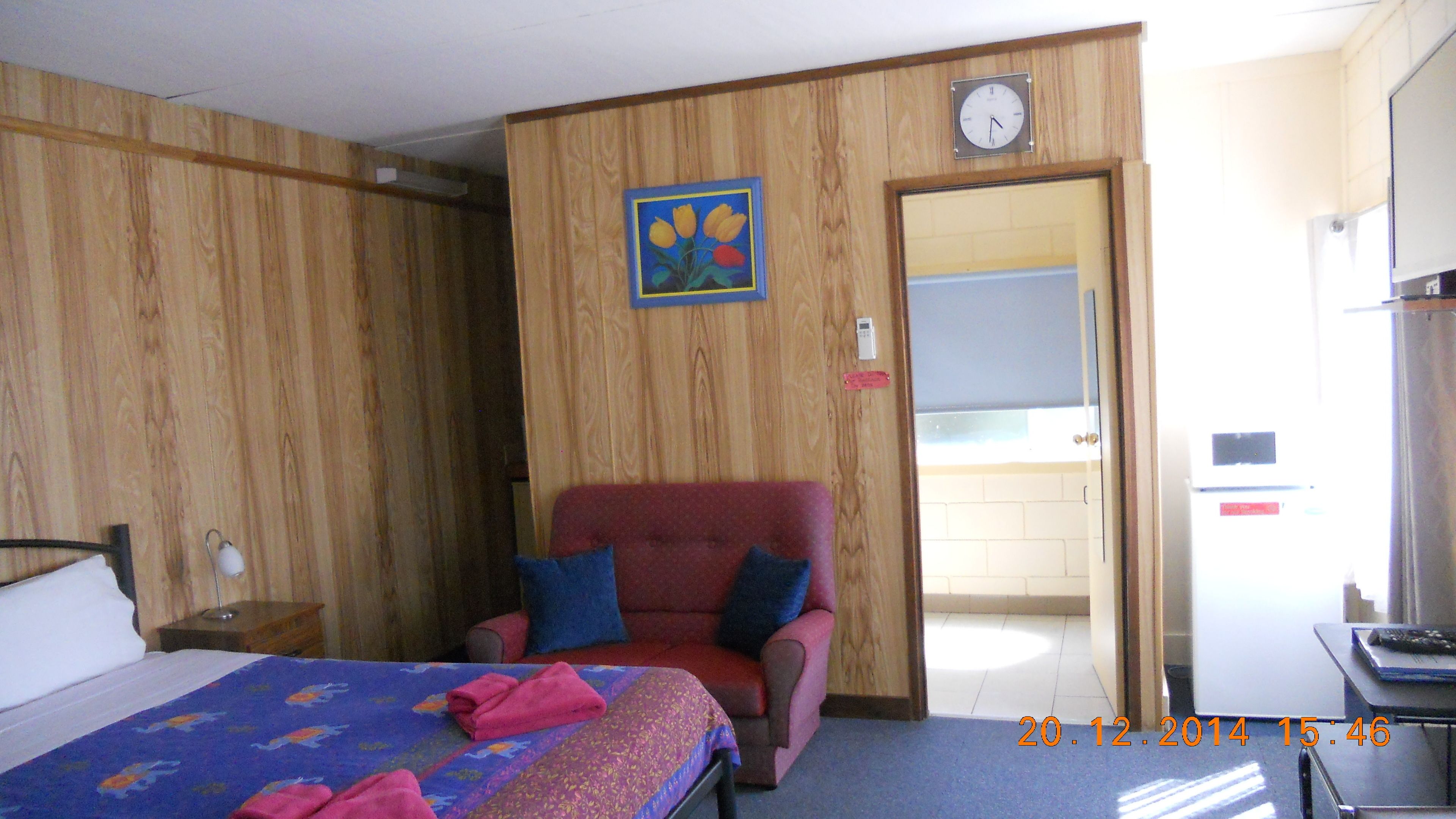 Quarto de hotel com uma cama, uma poltrona vermelha com almofadas azuis, uma parede de madeira, um relógio e uma porta que dá para outro quarto.