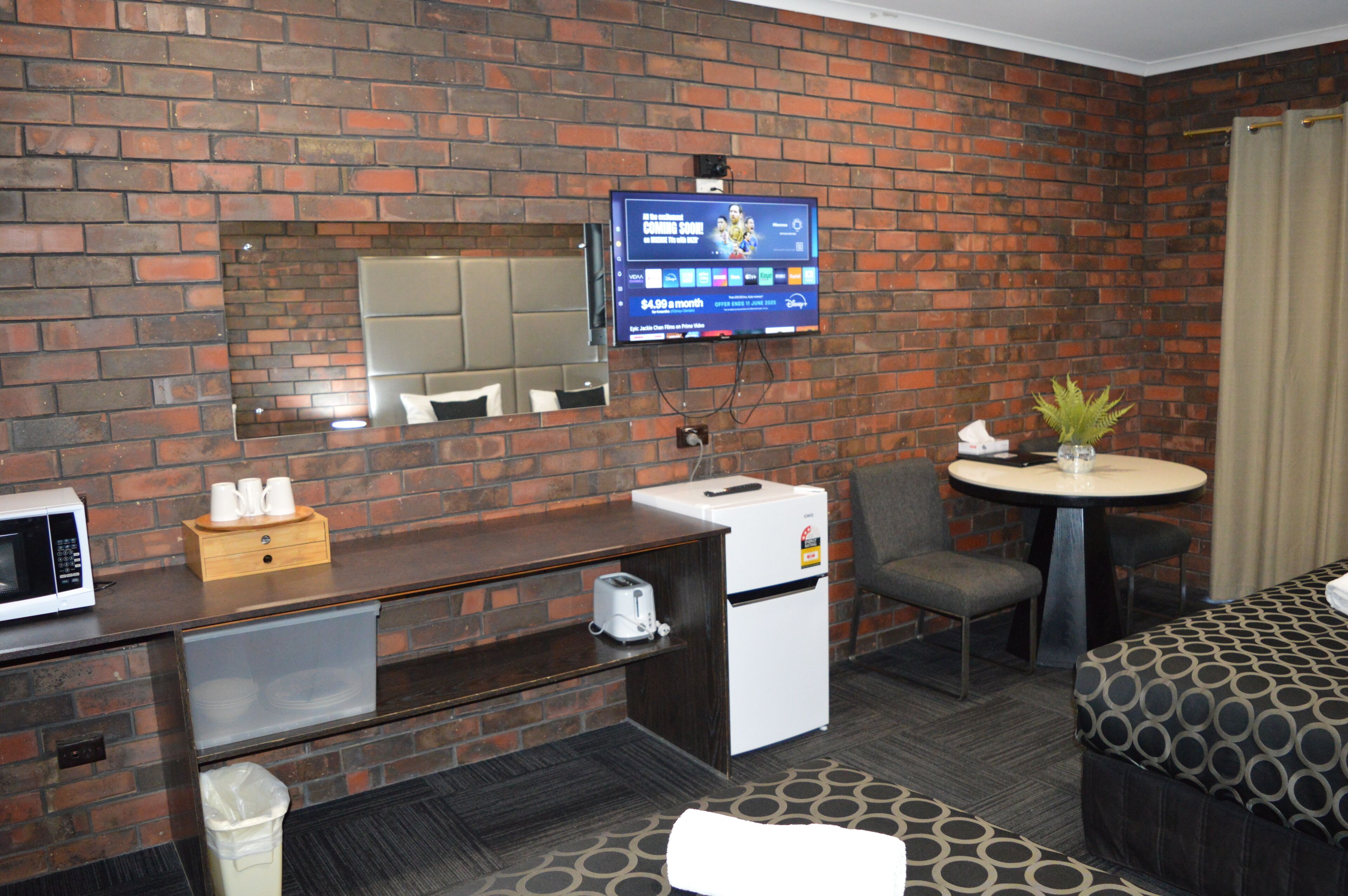A hotel room with a brick wall, a flat-screen TV mounted above a wooden desk, a small refrigerator, a microwave, a trash can, a round table with a potted plant, and a patterned sofa.
