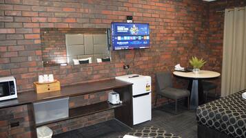 A hotel room with a brick wall, a flat-screen TV mounted above a wooden desk, a small refrigerator, a microwave, a trash can, a round table with a potted plant, and a patterned sofa.
