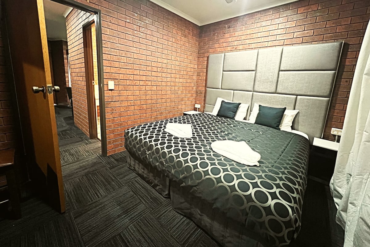A hotel room with a brick wall, a large bed with a patterned bedspread, a ceiling fan, and a nightstand with a lamp.