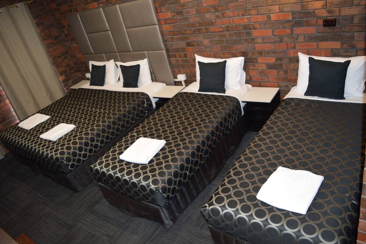 A hotel room with three single beds, each with a patterned bedspread and white pillows. The room has a brick wall and a grey headboard.