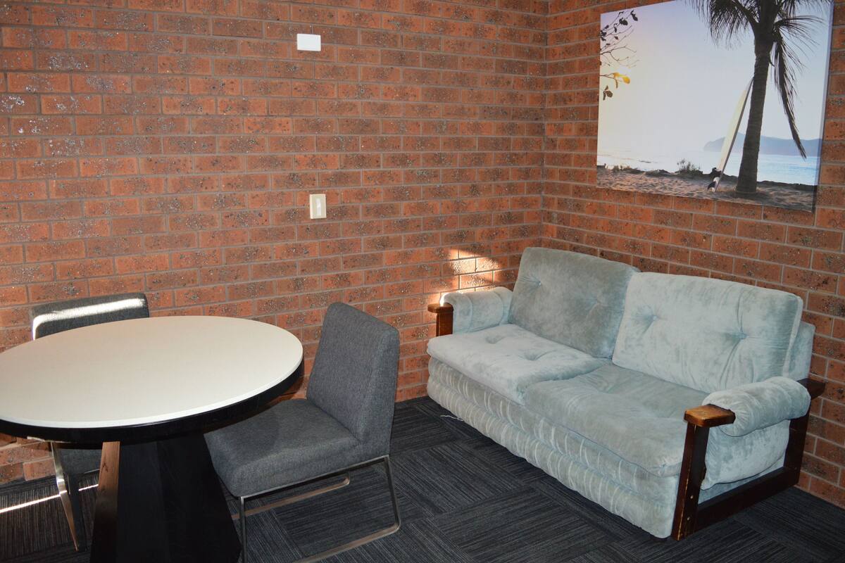 A room with a brick wall, a round table with a chair, a light blue sofa, and a picture of a beach scene.