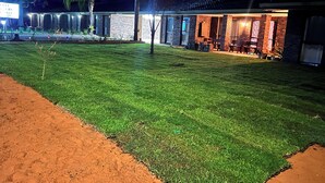 Un espace extérieur bien éclairé, avec une pelouse verdoyante, un bâtiment en briques doté d’un patio couvert et une enseigne indiquant le nom de la propriété.