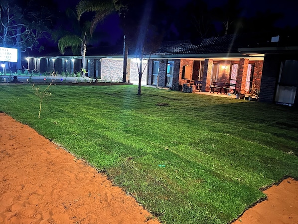 A well-lit outdoor area with a grassy lawn, a brick building with a covered patio, and a sign indicating the name of the property.