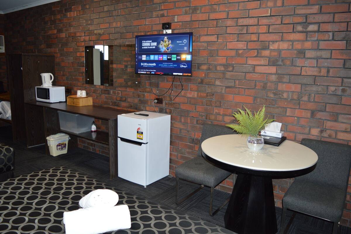 A hotel room with a brick wall, a flat-screen TV mounted above a minibar, a small refrigerator, a round table with a vase, and two chairs.