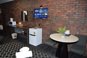 Et hotelværelse med en murstensvæg, et fladskærms-tv monteret over en minibar, et lille køleskab, et rundt bord med en vase og to stole.