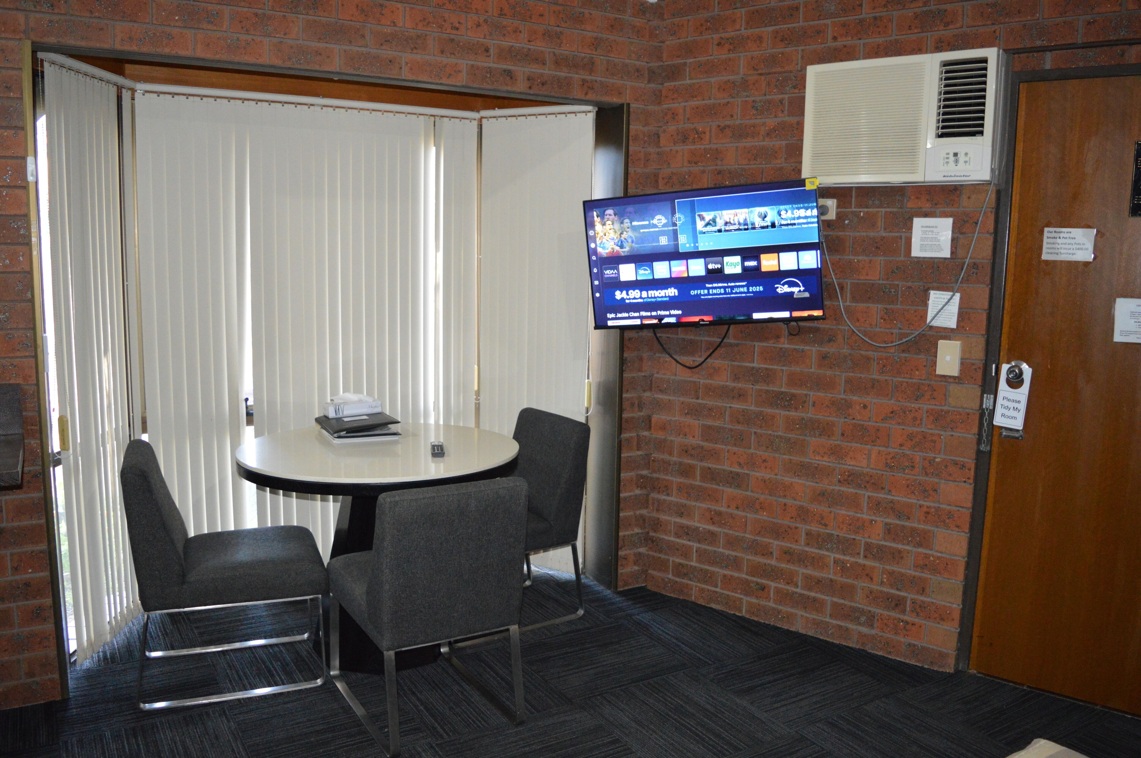 A conference room with a round table, four chairs, a flat-screen TV mounted on the wall, and a brick wall.