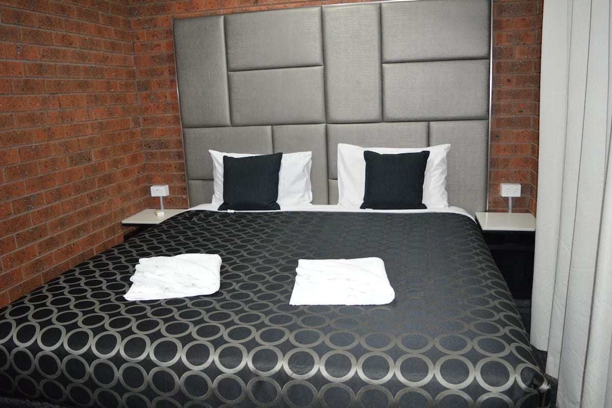 A hotel room with a brick wall, a grey padded headboard, two white pillows, a black and grey patterned bedspread, and two white towels on the bed.