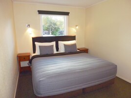 Quarto com cama, duas mesas de cabeceira, janela com persianas e duas luzes fixadas na parede.