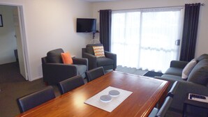 A modern living room with a wooden dining table, two grey sofas, a flat-screen TV mounted on the wall, and a large window with curtains. - Spa Lodge Motel (Hanmer Springs)
