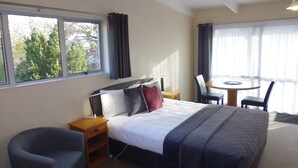 A hotel room with a bed, a chair, a small table, and a window with curtains. - Spa Lodge Motel (Hanmer Springs)