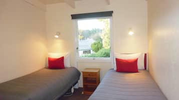 A small, well-lit room with two single beds, each with a red pillow, a small wooden nightstand, and a window providing a view of greenery outside.