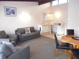 Una sala de estar con un sofá gris, una mesa de comedor con sillas, y una zona de cocina con microondas y fregadero.