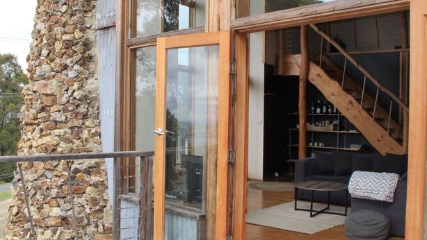Una terraza de madera con un muro de piedra, una puerta de cristal que da a una sala de estar con sofá y escalera.