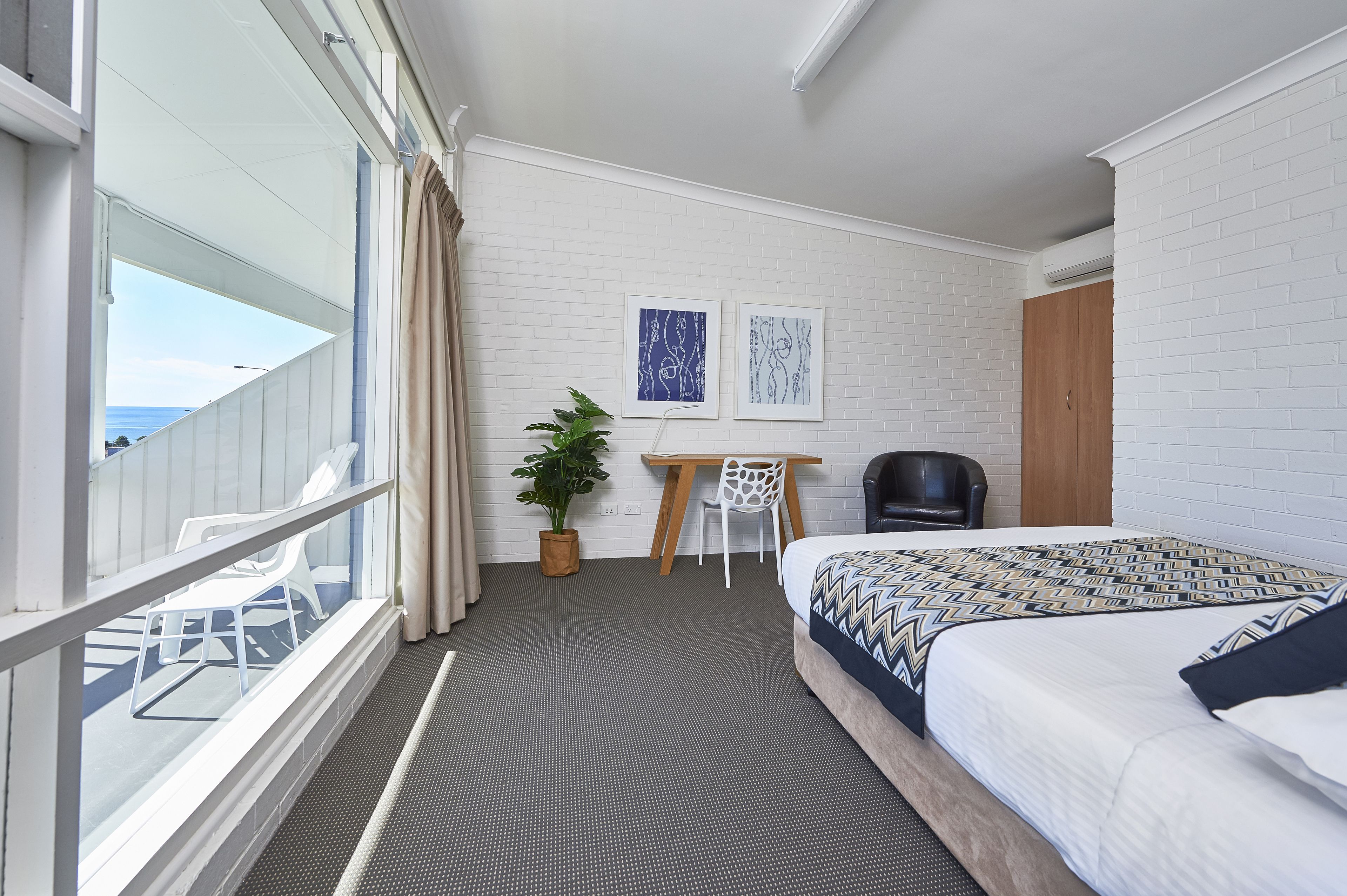 A hotel room with a balcony, a bed with patterned bedding, a desk with a chair, and two framed pictures on the wall.