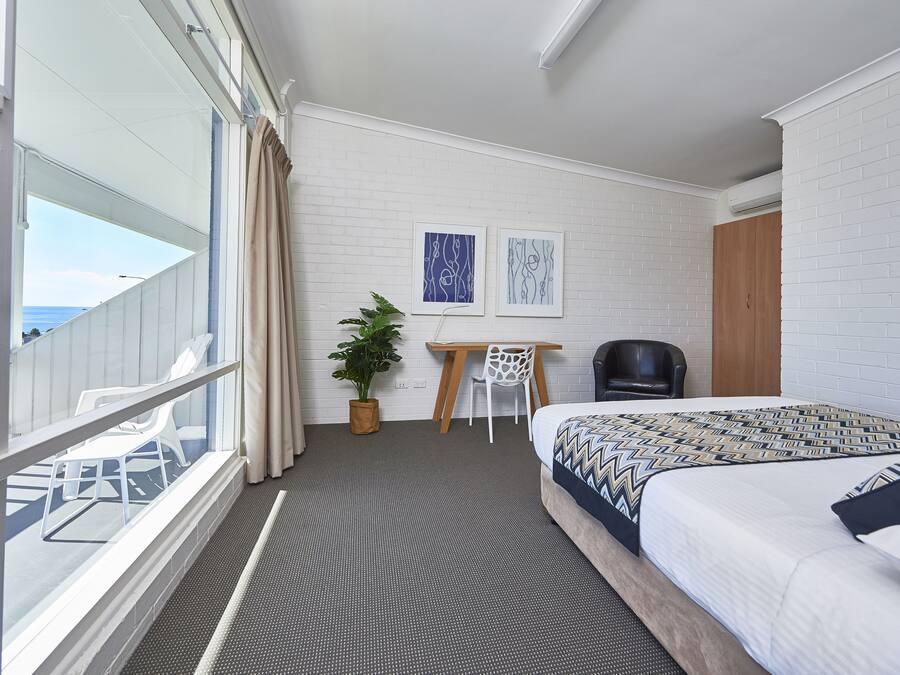 A hotel room with a balcony, a bed with patterned bedding, a desk with a chair, and two framed pictures on the wall.