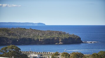 Paisaje costero con un promontorio rocoso, mar azul cristalino y una cadena montañosa en la distancia.