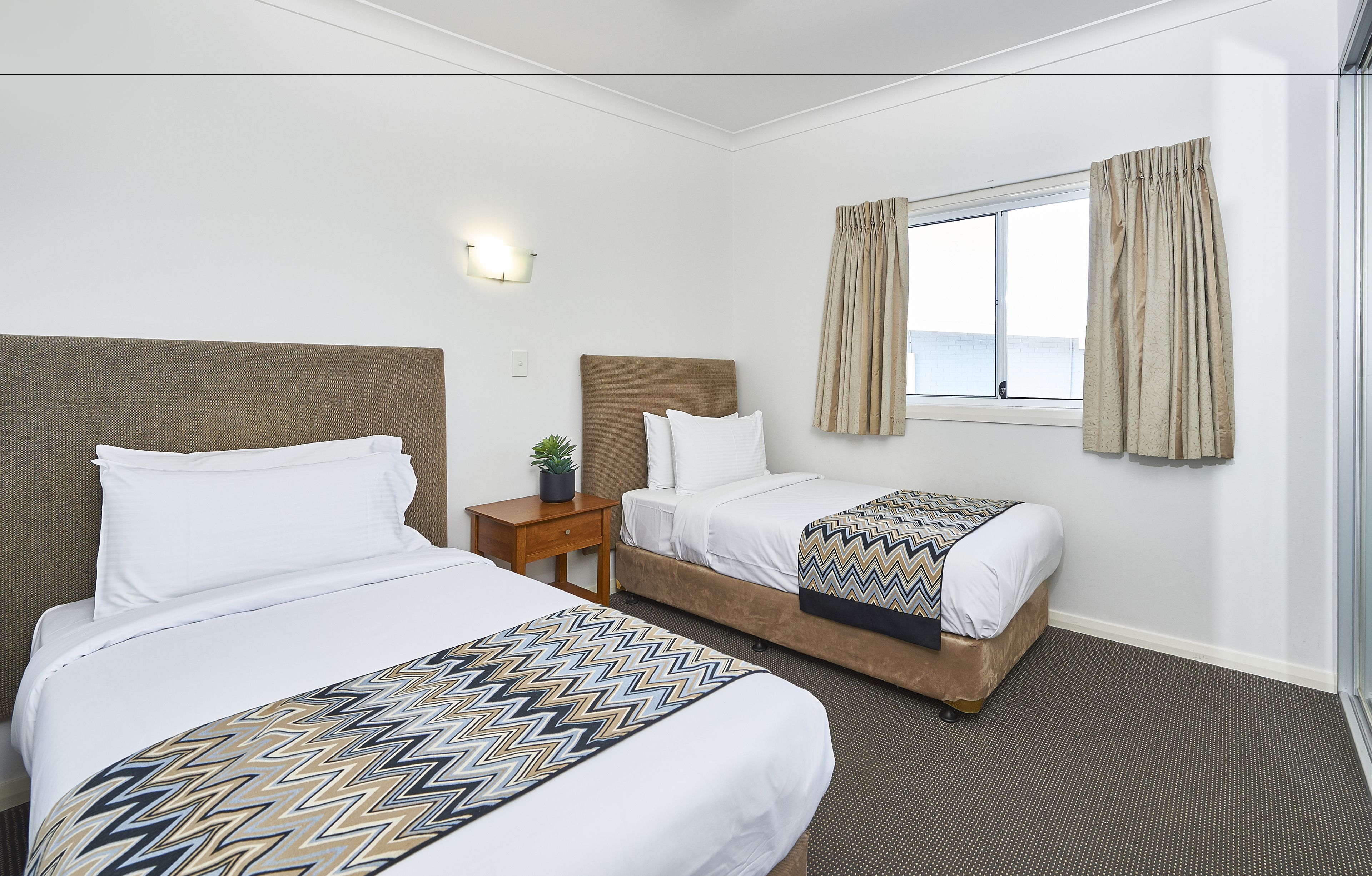 A hotel room with two beds, a nightstand, a potted plant, and a window with curtains.