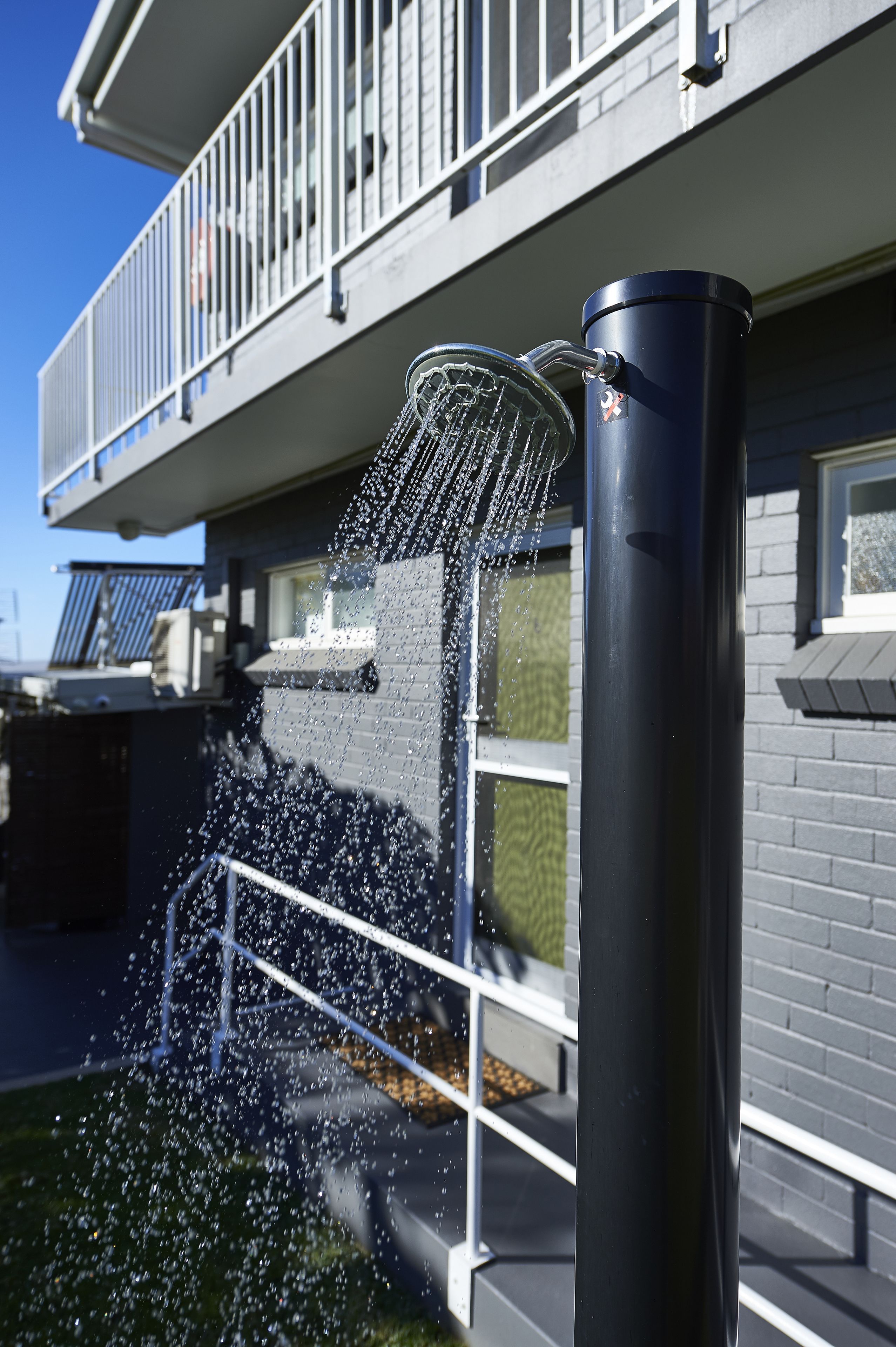 A water feature with a metal spout and a cylindrical base, set against a building with a balcony and windows.