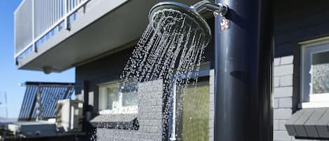 A water feature with a metal spout and a cylindrical base, set against a building with a balcony and windows.
