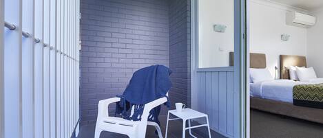 A balcony with a white plastic chair and a small table, a blue blanket on the chair, and a view of a bedroom with a bed and a nightstand.
