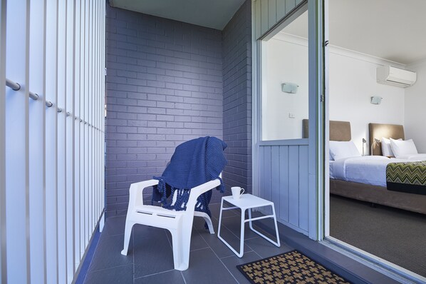 A balcony with a white plastic chair and a small table, a blue blanket on the chair, and a view of a bedroom with a bed and a nightstand.
