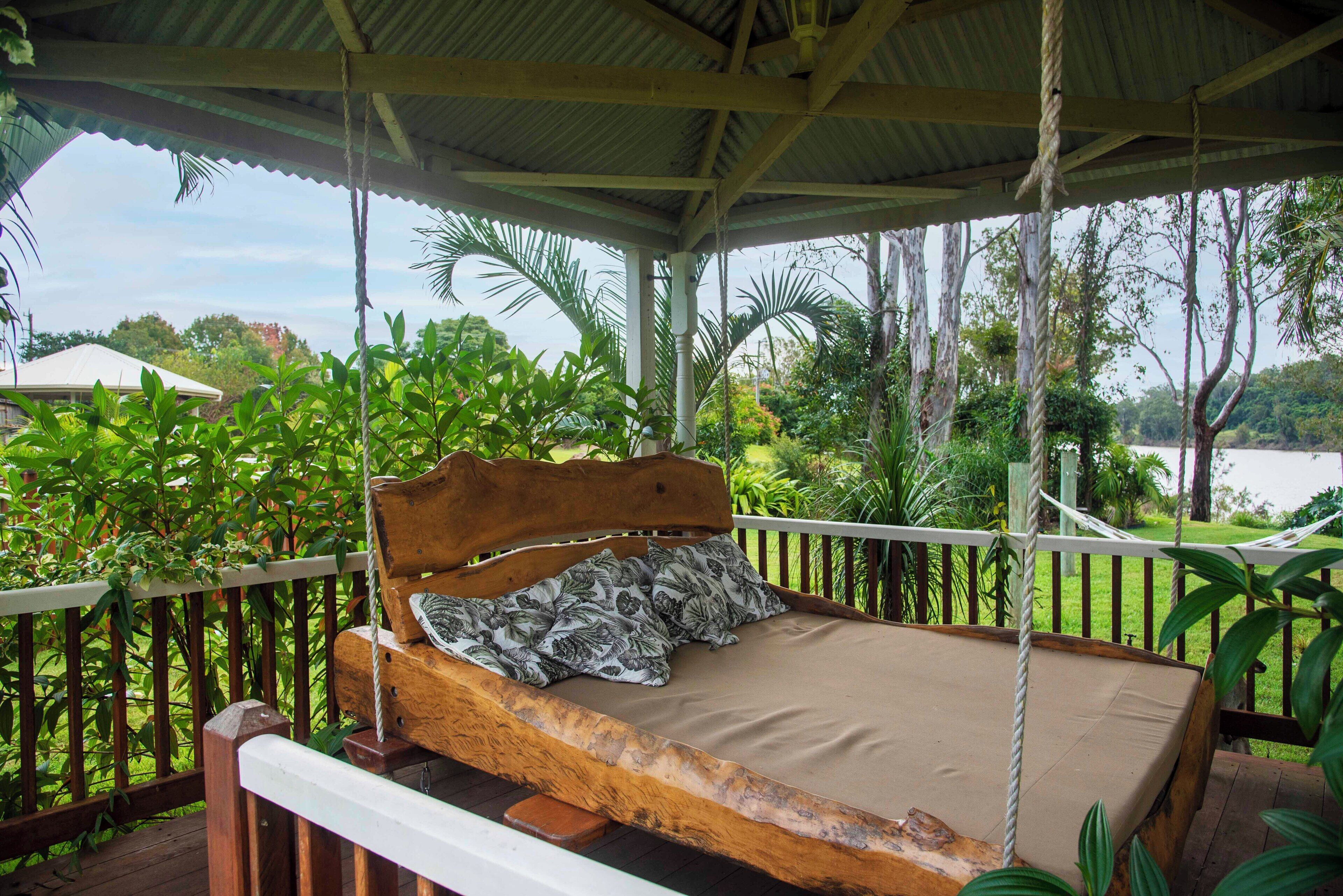 A wooden bed with a hammock-style headboard on a porch with a view of a lake and surrounding greenery.