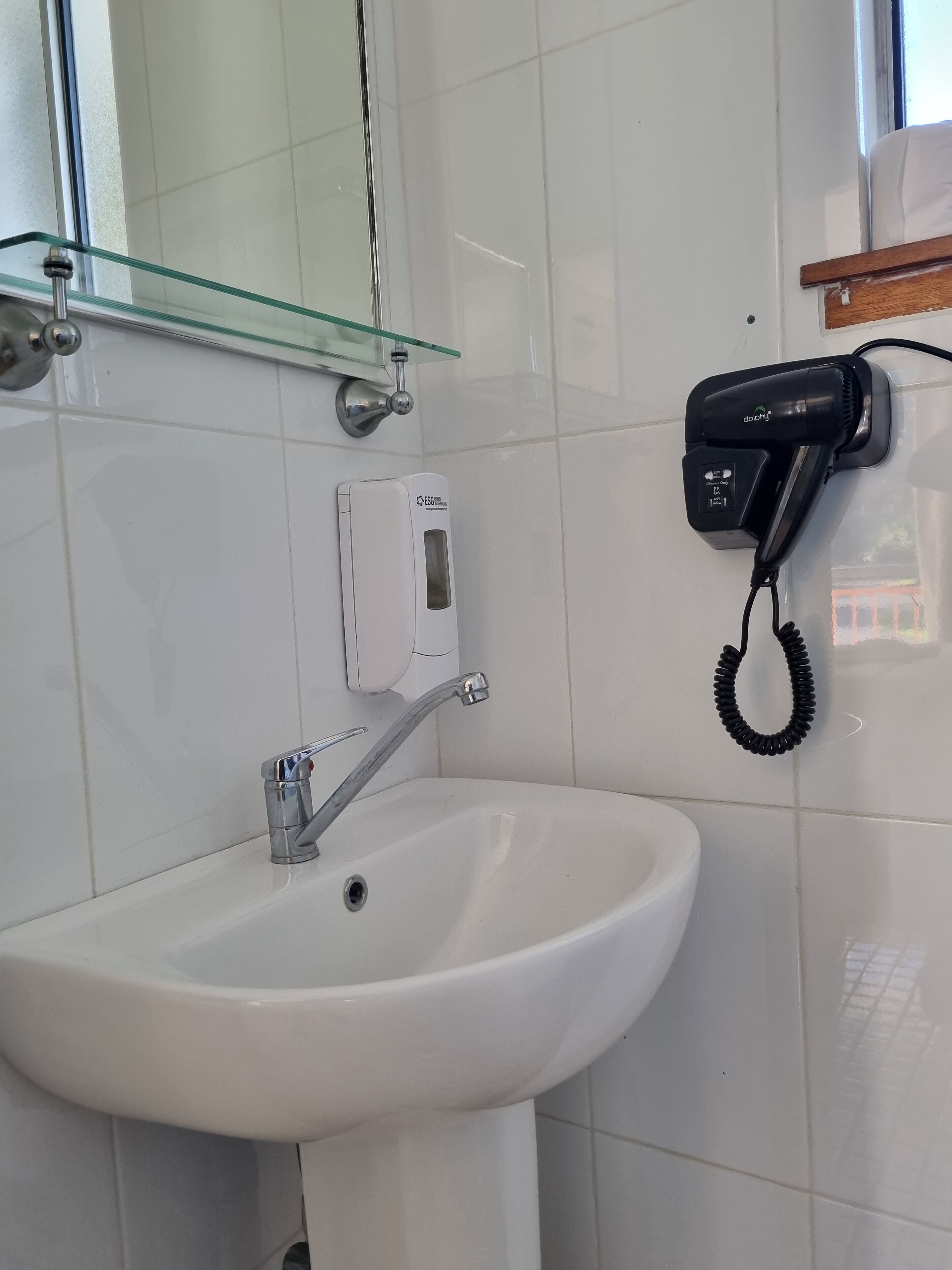 A bathroom with a white sink, a glass shelf, a hairdryer, and a soap dispenser.