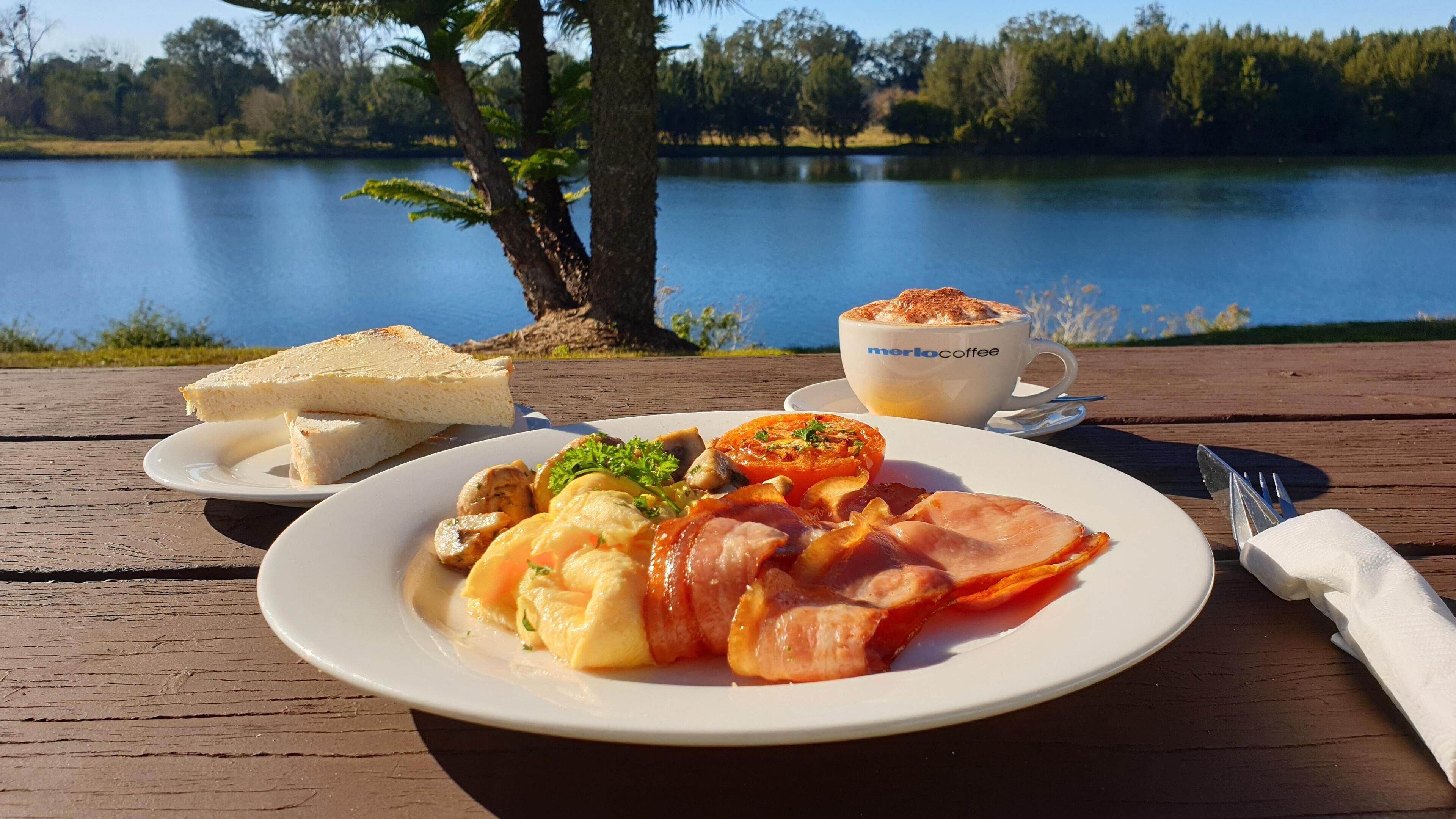 Een maaltijd op een houten tafel aan een meer, bestaande uit een bord met eieren, spek, champignons en een plak tomaat, een kop koffie en een sneetje brood.