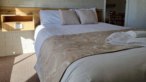 A neatly made bed with a patterned bedspread, a wooden shelf above it, and a power outlet on the wall. - Moon River Motor Inn (Kempsey)