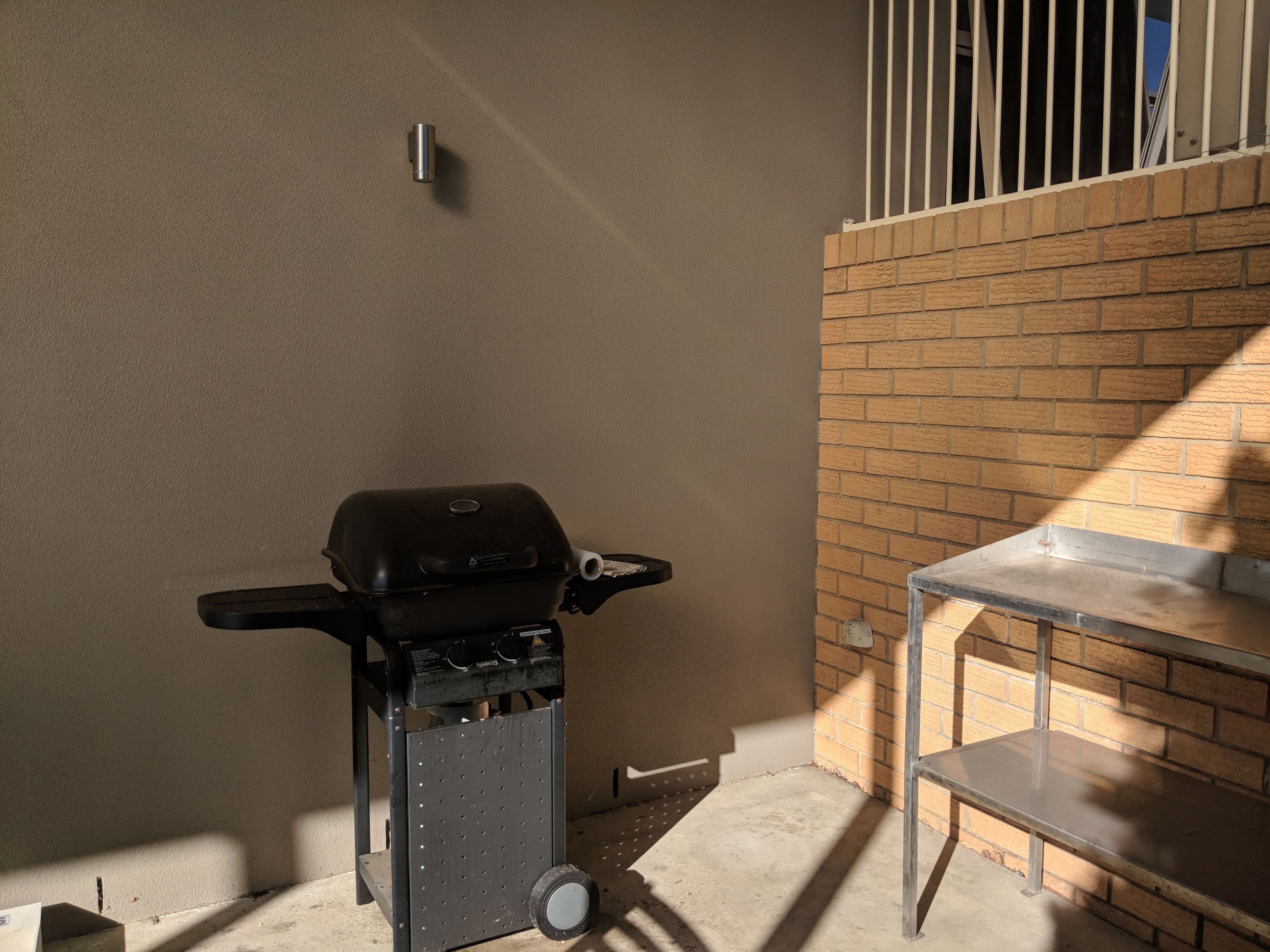 Una parrilla portátil en un rincón, con un muro de ladrillo y una baranda de metal.
