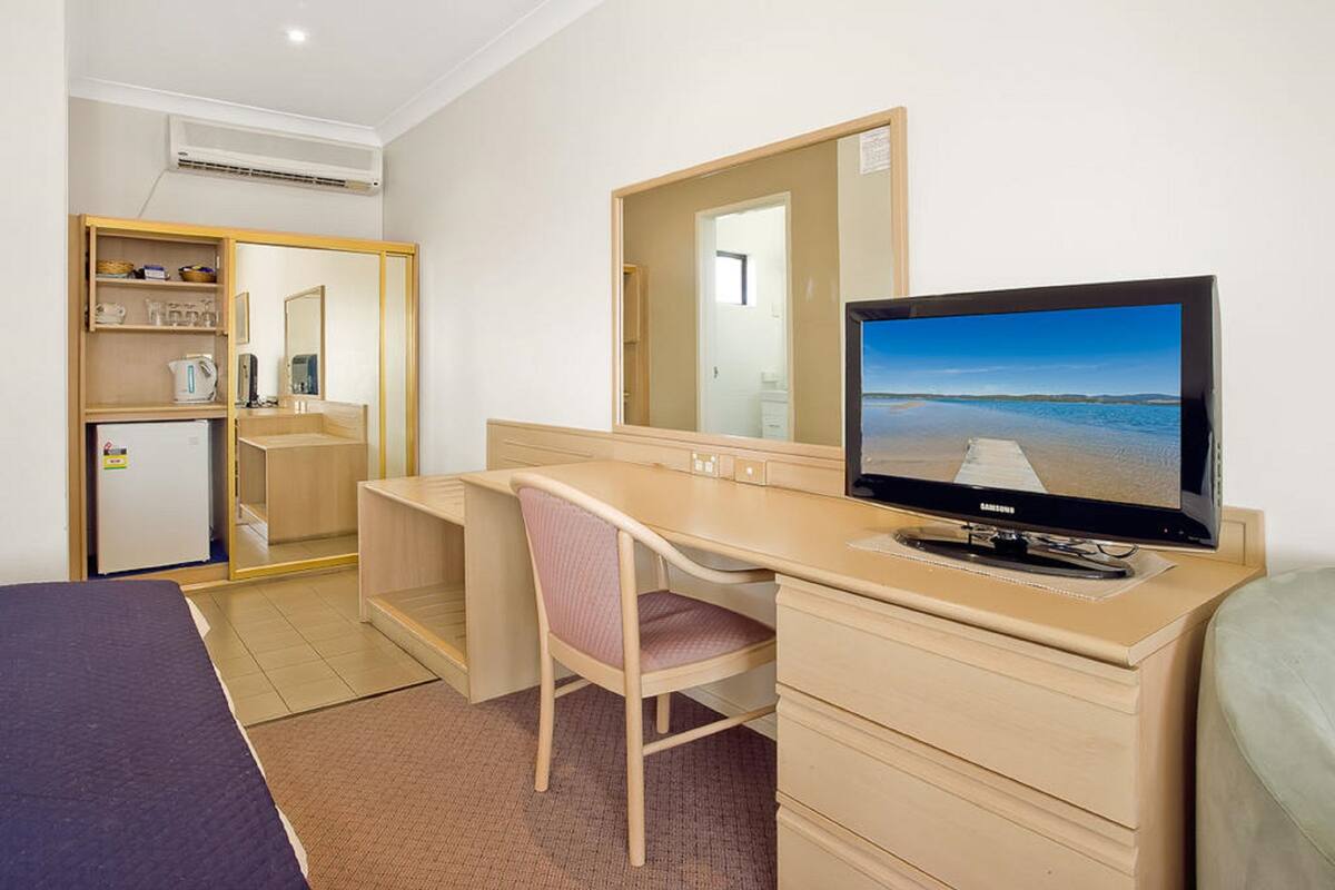 A hotel room with a bed, a desk with a chair, a television, a wardrobe, and a mirror.