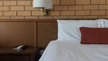 A hotel room with a brick wall, a wooden headboard, a bed with white and red pillows, a striped blanket, a nightstand with a lamp, and a drawer with a remote control.