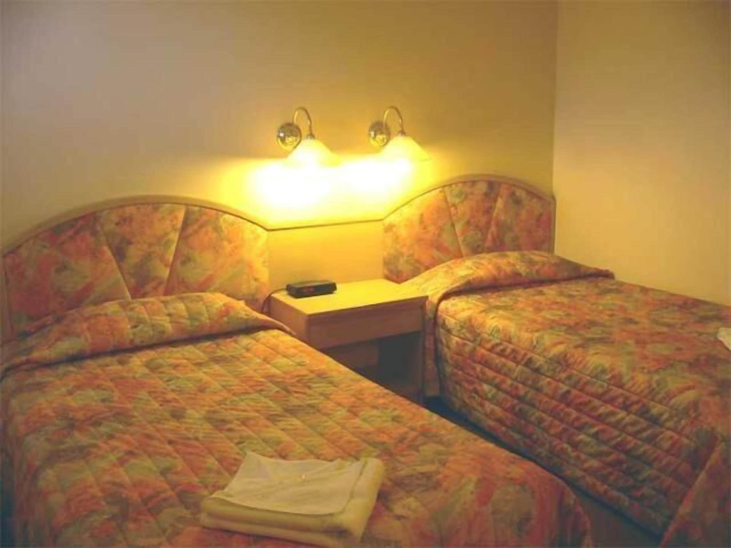 Hotellihuone, jossa on kaksi erillistä sänkyä, yöpöytä ja lamppu.