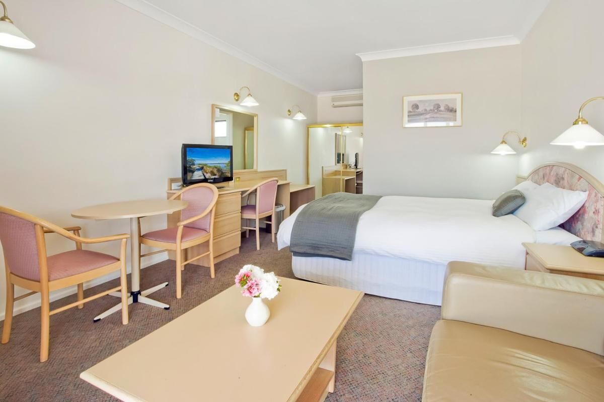 Habitación de hotel con una cama, un escritorio con silla, una mesita con un jarrón de flores y un televisor.