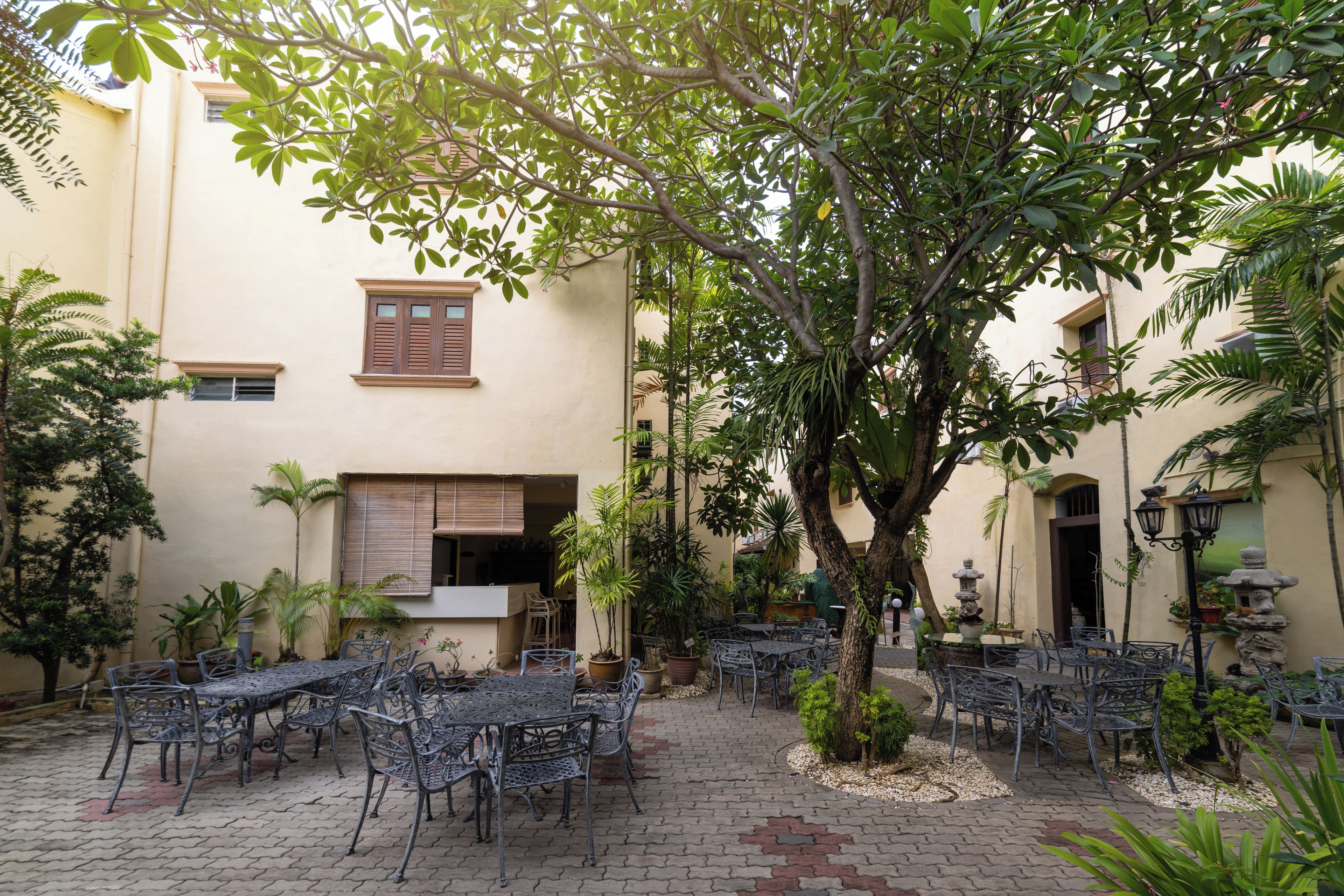 An outdoor patio with metal tables and chairs, surrounded by greenery and a building with windows and shutters.