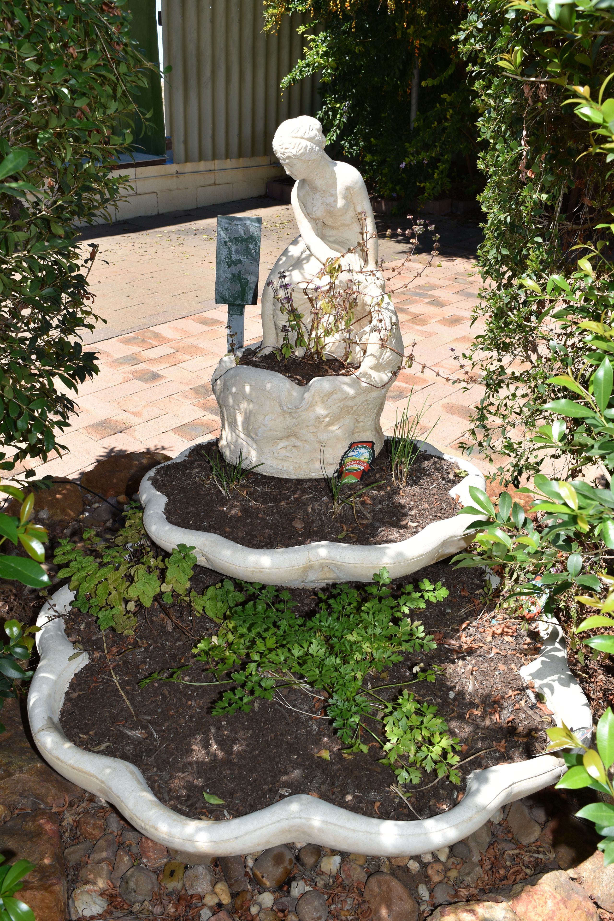 Una statua da giardino raffigura una figura seduta con una pianta sulle gambe, circondata da un bordo bianco e un piccolo vaso con terra e piante.
